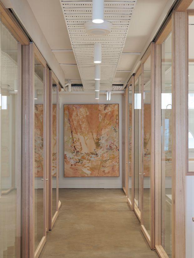 A refined interior corridor lined with full-height glazed timber-framed partitions and doors, creating a series of office or gallery-like rooms on either side. The passage has a polished concrete-toned floor, pale walls, a suspended perforated acoustic ceiling with cylindrical pendant lights, and a large abstract artwork in warm earthy tones at the end.