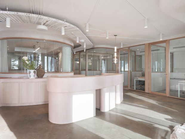 A bright contemporary commercial interior with a large curved pale reception or service counter in the foreground, surrounded by curved glazed partitions and timber-framed glass offices. The space features a polished concrete floor, white ceiling with exposed linear detailing and track lighting, pale timber cabinetry, and abundant natural light.