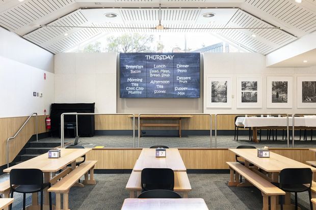 Bright, modern multi-level dining or hospitality space with tiered timber seating platforms, long communal tables, black chairs, a central menu display board, framed artwork, and extensive white acoustic ceiling panelling. Metal balustrades and accessible handrails define the raised platforms.