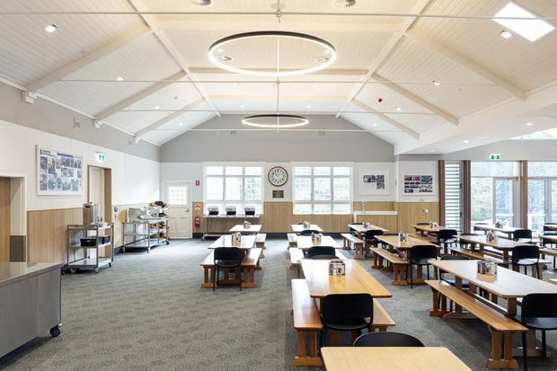 A bright, spacious institutional dining hall or cafeteria with rows of timber tables and black chairs, a carpeted floor, serving trolleys, and large multi-pane windows. The room has a high white pitched ceiling with exposed beams, circular pendant lights, white walls, and light timber wall panelling. Posters and noticeboards line the walls, while daylight enters through the windows and glazed doors.
