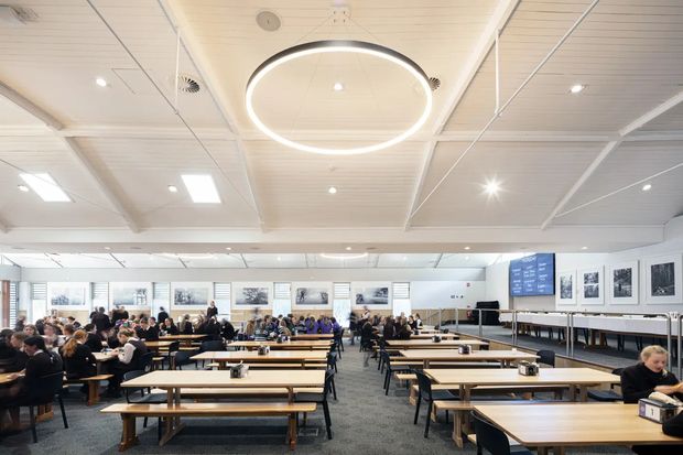 A large, bright institutional dining hall or cafeteria with numerous long timber tables and bench seats arranged in rows. The space has a white paneled suspended ceiling with recessed lighting and a large circular pendant light, grey carpeted flooring, framed artworks along the walls, and extensive glazing at the far end. People are seated throughout the room.