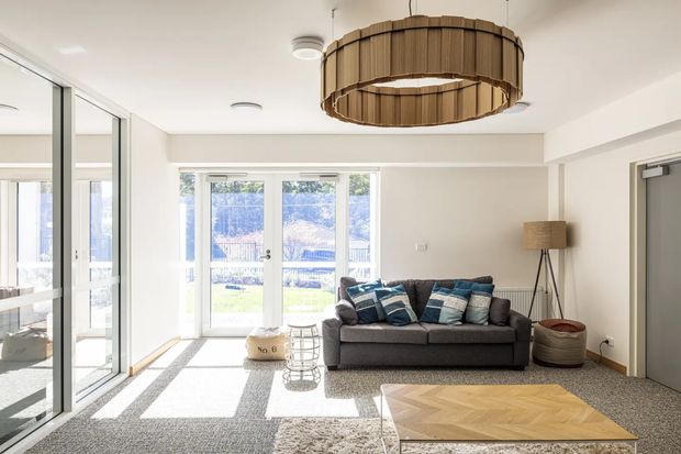 Bright contemporary living room with white walls, large glazed doors and windows opening to a garden, grey carpet flooring, a dark grey sofa with blue and neutral cushions, and a prominent suspended circular timber-slat pendant light. A light timber herringbone-pattern coffee table and tripod floor lamp complete the space.