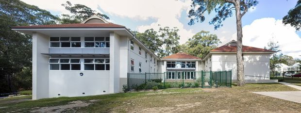 Exterior view of a low-rise institutional or commercial building complex with white rendered walls, red tiled hipped roofs, horizontal window bands, a large enclosed opening on the left building, green metal fencing, paved walkways, and mature trees surrounding the site.