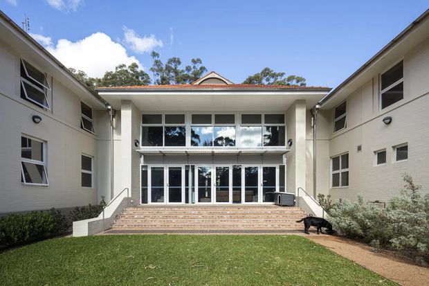 Symmetrical exterior view of a low-rise institutional or residential building arranged around a central entry courtyard. The cream rendered wings frame a projecting central entrance with broad tiled steps, full-height glazed doors, a deep flat-roof canopy, and an upper-level glazed balcony or window wall. A small gabled roof element is visible behind the central canopy. The scene includes a lawn, simple garden planting, and a clear blue sky.