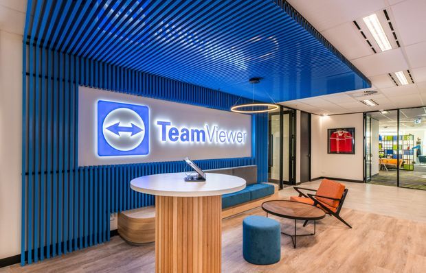 A contemporary commercial reception and breakout lounge featuring a prominent illuminated TeamViewer logo, a blue slatted feature wall and ceiling, curved timber reception desk, blue upholstered bench and ottoman, lounge chairs, coffee table, glass partitions, and light timber-look flooring.
