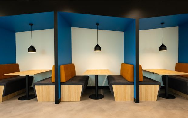 A row of compact booth-style dining or meeting alcoves separated by bold blue structural partitions. Each booth has a light timber table, upholstered orange and dark grey bench seating, black pedestal bases, and a black pendant light against a white wall. The space has a polished concrete floor and a dark ceiling, creating a contemporary commercial interior with strong colour contrast.