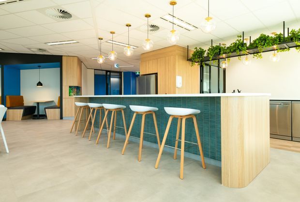 A bright contemporary commercial breakout kitchen and dining area with a large island bench, white benchtop, vertically fluted light-wood cabinetry, and a teal small-format tiled island face. White stools with timber legs line the island. Pendant globe lights hang above, while open shelving with trailing greenery adds a natural accent. Blue feature walls and glazed meeting-room partitions are visible in the background beneath a suspended acoustic ceiling.