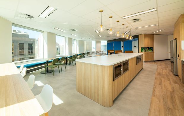 Bright contemporary commercial office breakout and kitchenette area with a large light-wood island, integrated appliances, white benchtops, pendant lighting, communal dining tables, and extensive windows. Blue accent joinery and timber flooring add colour and warmth to the open-plan workspace.