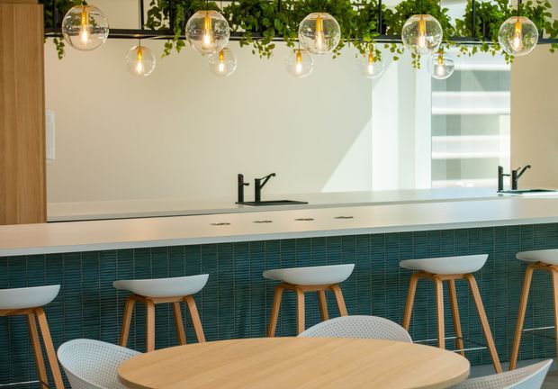 Bright contemporary kitchen or hospitality-style bar area with a long white counter, multiple black faucets, teal textured tile facing, timber-legged white stools, and a light timber dining table with white perforated chairs. Globe pendant lights and a hanging greenery feature add visual interest, while a large mirror reflects the counter and joinery.