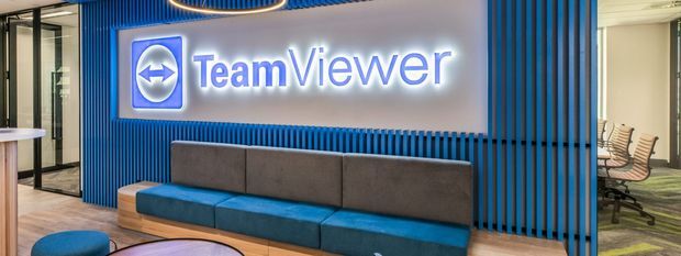 A contemporary commercial office reception or lounge area featuring a prominent illuminated TeamViewer logo mounted on a grey feature wall. Vertical blue slatted wall panels frame the signage, with a long blue-and-grey upholstered built-in sofa, timber flooring, glass partitions, office chairs, and a circular pendant light.