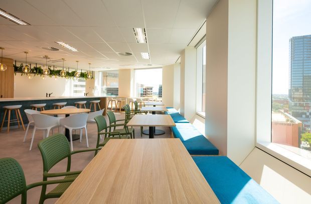 Bright contemporary commercial dining and breakout area with extensive floor-to-ceiling windows, light timber tables, green chairs, and a long vivid blue upholstered window bench. A compact bar or kitchenette with pendant lighting and hanging greenery is visible at the rear, with an urban office or city view beyond the glazing.