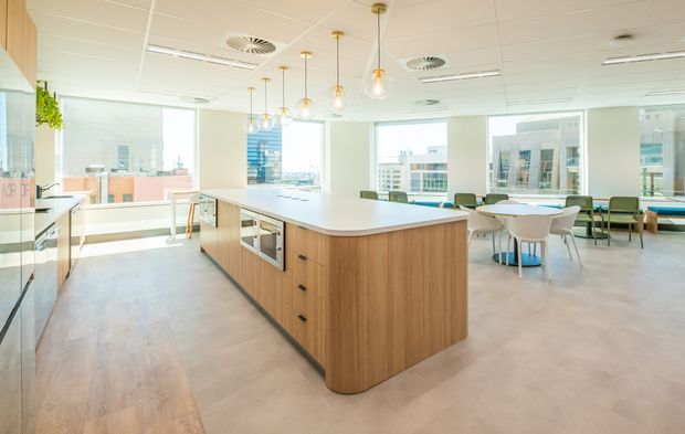Bright contemporary commercial kitchenette and breakout area with a large central timber island, integrated appliances, pale flooring, pendant globe lights, and casual dining tables beside expansive city-facing windows.