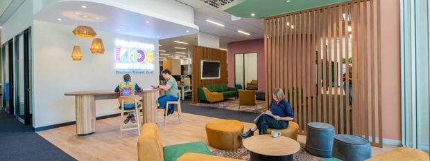 A bright, contemporary communal interior with a casual meeting and lounge area. The space features pale timber-look flooring, white walls, a curved feature ceiling with recessed lighting, pendant lights, a long high table with stools, colourful lounge seating, low round tables, and a prominent vertical timber slat screen dividing the room. People are using the space, with a projected presentation visible in the background.