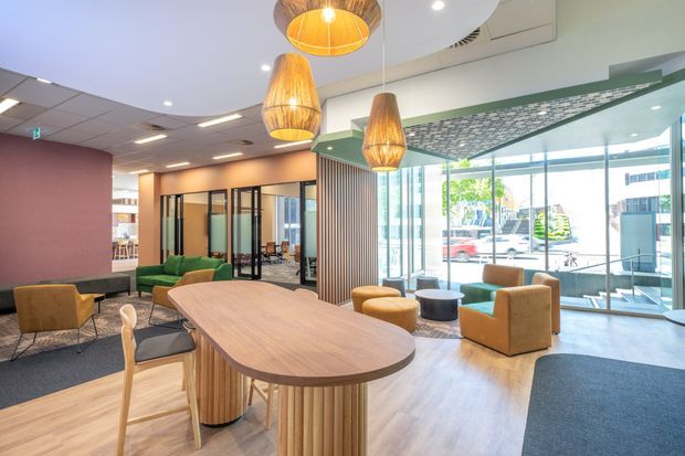 Contemporary commercial office lounge and collaboration area with a large rounded timber communal table, green and mustard upholstered seating, glass meeting-room partitions, and floor-to-ceiling glazing. Feature timber pendant lights, vertical timber slat detailing, a pale timber-look floor, and a patterned ceiling transition create a warm, welcoming workplace interior.