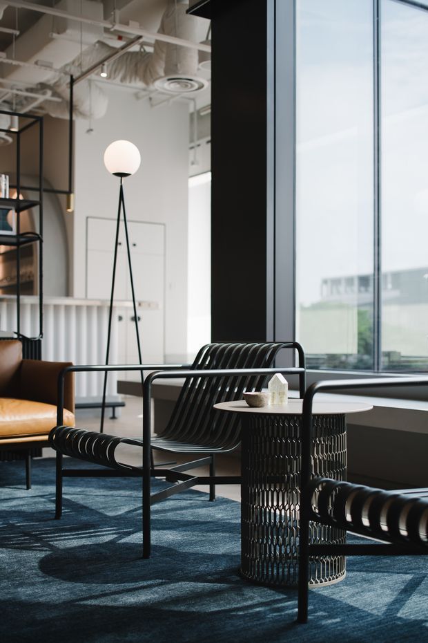 A stylish contemporary lounge or reception area with floor-to-ceiling glazing, dark blue carpet, black metal-framed lounge chairs, a round black wire-base side table, and a warm brown leather armchair. A sculptural tripod floor lamp, open shelving, and exposed white ceiling services contribute to an industrial-inspired commercial interior.