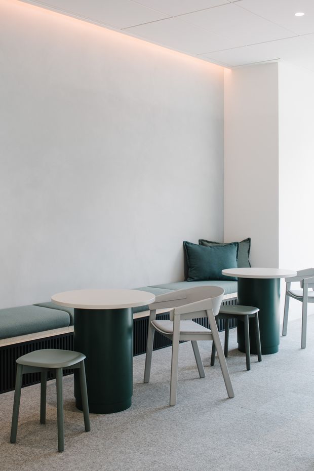 A minimalist, softly lit commercial interior with pale walls, a white suspended ceiling, and grey carpet. A built-in upholstered bench runs along the wall beneath concealed warm cove lighting, paired with small round tables, dark green cylindrical bases, stools, and light timber-framed chairs. The space appears to be a contemporary café, lounge, or informal breakout area.