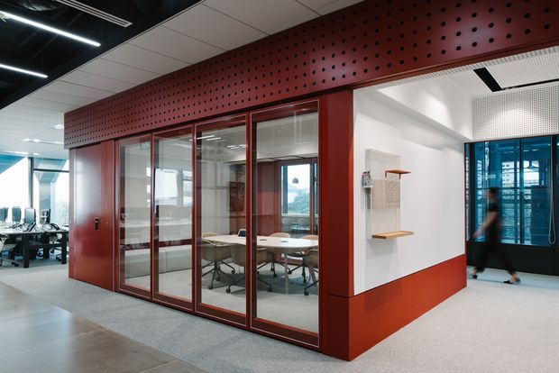 A contemporary commercial office interior featuring a glazed meeting room enclosed by dark red metal-framed glass partitions. The meeting room has a conference table and office chairs, while the surrounding space includes open work areas, exposed black ceiling elements, linear lighting, white acoustic ceiling panels, and perforated red feature cladding above the enclosure. A small built-in shelf and coat storage are visible on the adjoining white wall.