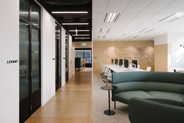 A contemporary commercial office interior with a reception or breakout lounge in the foreground, featuring a large curved dark green upholstered sofa, small round side tables, and a long communal dining or meeting table with white chairs. Glazed black-framed meeting-room doors line the left side, while a perforated timber-toned partition and botanical line-art wall graphic define the rear. The space has polished concrete or terrazzo-like flooring, a suspended white acoustic ceiling with linear recessed lighting, and exposed dark services in the corridor ceiling.