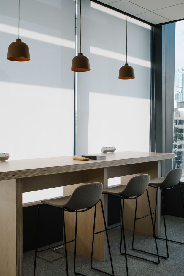 A minimalist contemporary commercial workspace or breakout area with a long light-wood communal counter, integrated power outlets, and several dark upholstered bar stools. Three suspended dark pendant lights hang in front of large windows with roller blinds, while a city building is visible through the glazing. The space has a clean, restrained palette of white, light wood, charcoal, and grey.
