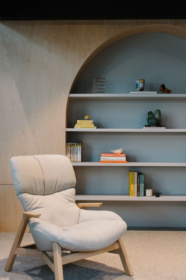 A softly lit reading nook featuring a light upholstered lounge chair with timber arms and legs, positioned in front of a built-in arched bookshelf. The shelving has a muted blue-grey painted backing, pale horizontal shelves, and a pale timber surround and wall panelling, with books and small decorative objects arranged throughout. A neutral carpet and textured rug complete the calm, curated interior.