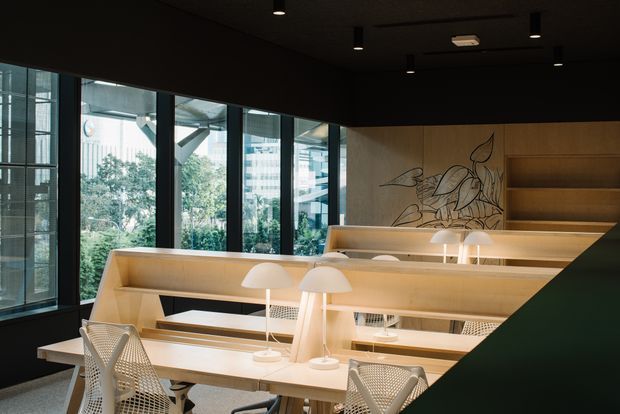 A refined, open-plan commercial workspace with tiered light-wood desk and workstation joinery arranged beside expansive floor-to-ceiling glazing. The space has a dark ceiling and walls, integrated shelving, warm desk lamps, mesh office chairs, and a decorative botanical line-art feature on the timber rear wall. Views through the glazing show an urban setting and greenery outside.