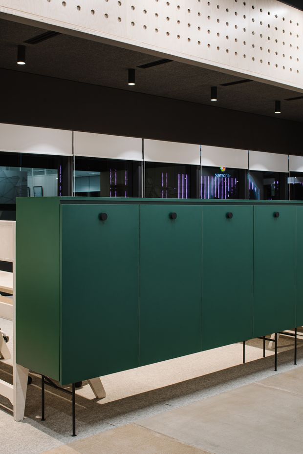 A contemporary commercial interior featuring a large dark green fixed storage or partition unit raised on slender black metal legs. The unit has flat panel doors with small round dark pulls. Behind it are glazed partitions and a dark ceiling with recessed linear elements and cylindrical downlights. Above, a pale perforated timber or wood-panel feature adds texture. The space has polished concrete flooring and a restrained, modern palette of deep green, black, pale wood, and grey.