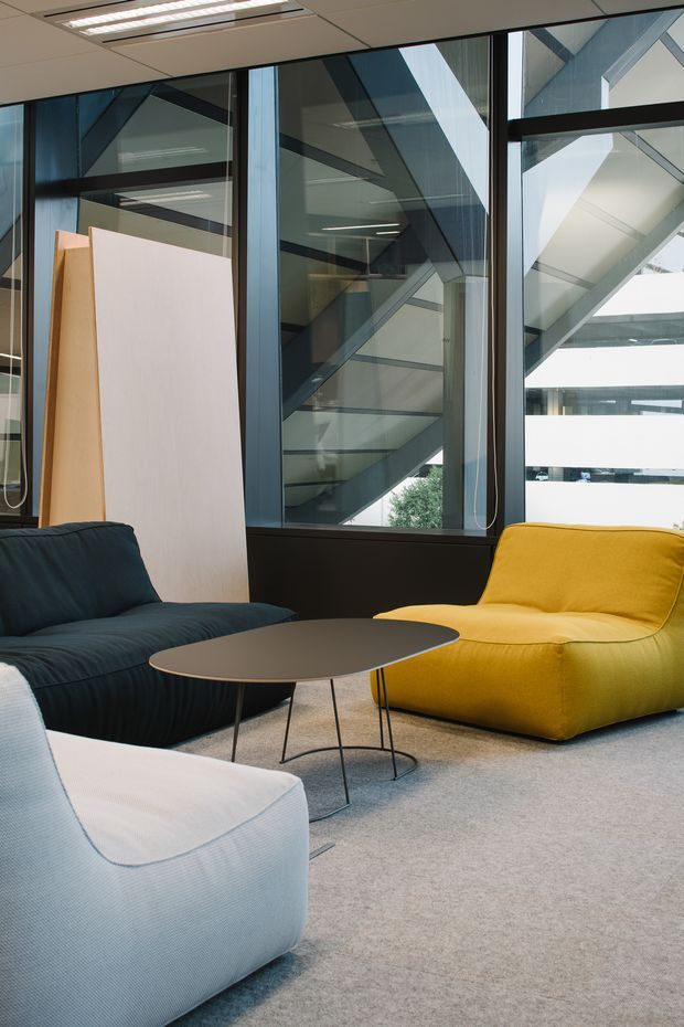 A contemporary lounge seating area with dark teal, yellow, and white upholstered lounge chairs arranged around a low oval coffee table. The space has pale carpet, dark-framed floor-to-ceiling glazing, and exposed structural elements and staircases visible beyond the glass, creating a bright office or commercial interior setting.