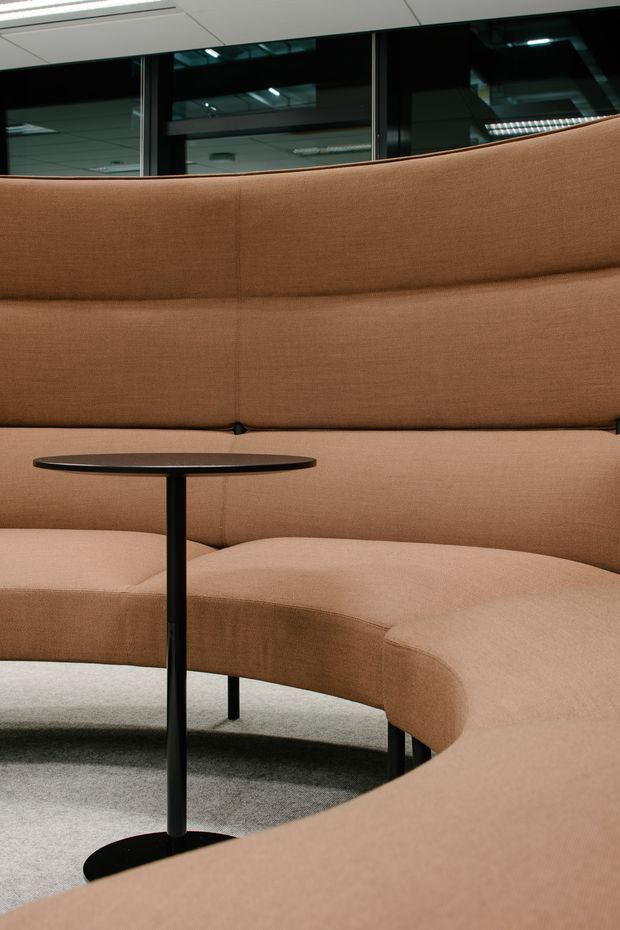 Close-up view of a curved upholstered banquette in a commercial interior, upholstered in warm tan fabric with visible stitched panels. A small black round pedestal table sits in front, on grey carpet. Dark-framed interior glazing and a suspended ceiling are visible behind the seating, suggesting a contemporary office or hospitality setting.