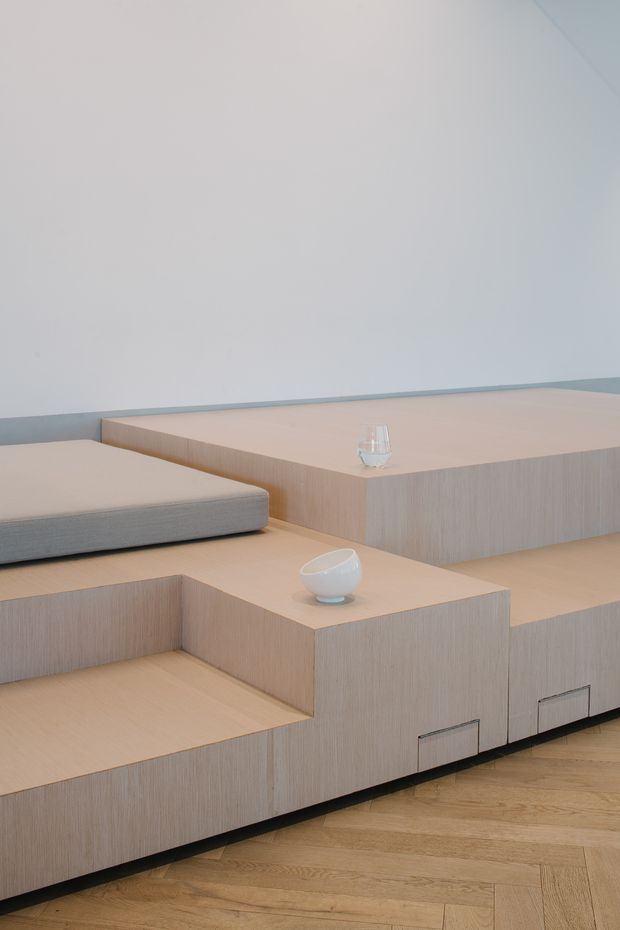 Minimalist interior seating area with tiered, pale timber built-in joinery forming stepped benches and low storage drawers. A light upholstered cushion and small ceramic and glass objects sit on the platforms. The room has smooth white walls, a narrow grey horizontal trim, and a warm timber herringbone floor.