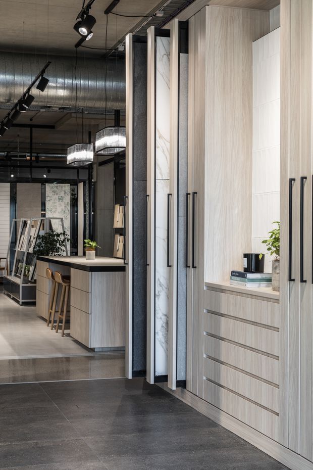A sophisticated contemporary kitchen showroom or kitchen interior featuring tall light timber-look cabinetry with long black handles, integrated drawers, white tiled and marble-look feature panels, and dark tiled flooring. A compact island with stools sits in the adjoining area, while exposed metal ductwork, black track lighting, pendant lights, and framed glass partitions create an industrial commercial atmosphere.