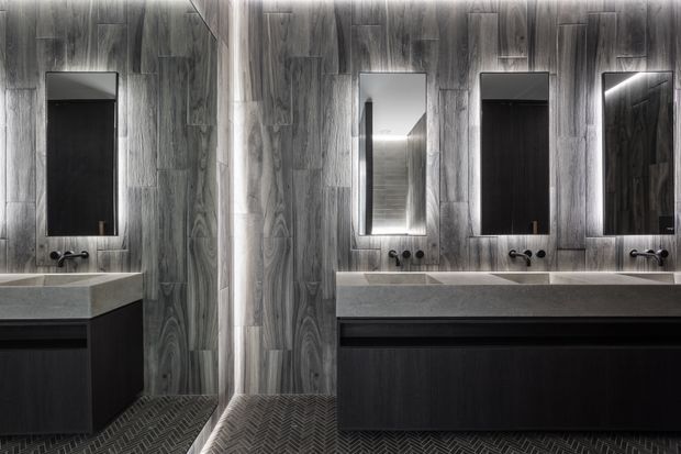 A dark, sophisticated bathroom with a long double vanity, multiple rectangular mirrors, black wall-mounted faucets, dark timber-look wall panelling, grey stone benchtops, and a charcoal herringbone tile floor. Vertical integrated lighting highlights the mirrors and vanity.