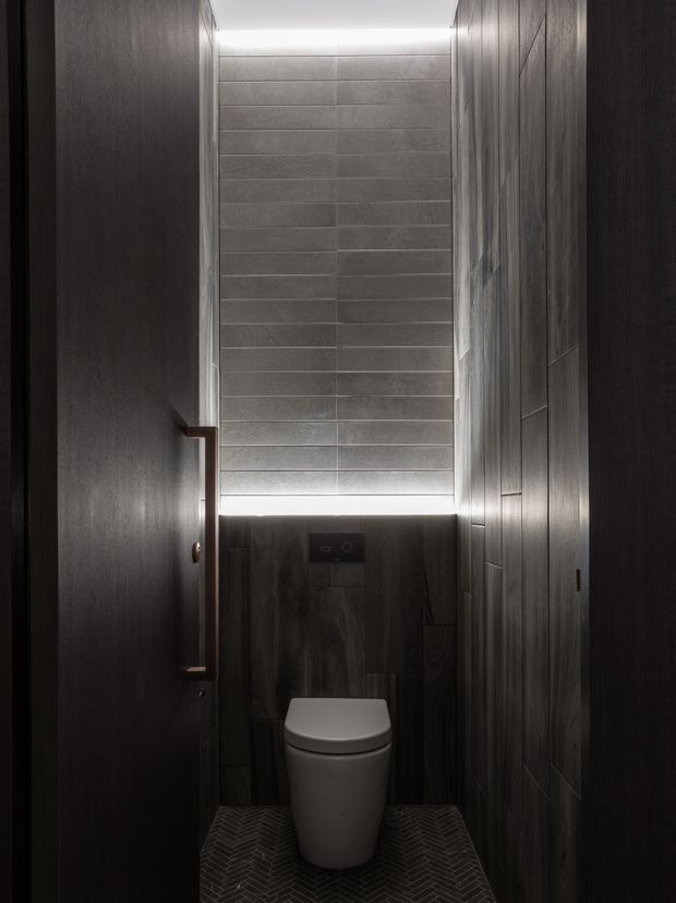 A narrow, dark contemporary toilet room with a wall-mounted toilet, charcoal wood-look wall panels, a grey tiled feature wall, and a dark herringbone tile floor. Concealed linear lighting illuminates the upper wall and ceiling edges, creating a dramatic, refined atmosphere. A partially visible timber door with a long vertical handle opens into the space.