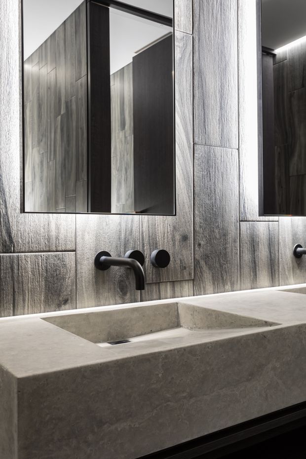 A refined contemporary bathroom vanity with a substantial double concrete trough basin, wall-mounted matte black faucets, large frameless mirrors, and dark vertically grained wall tiles or slabs. Integrated linear lighting adds a dramatic, moody atmosphere.