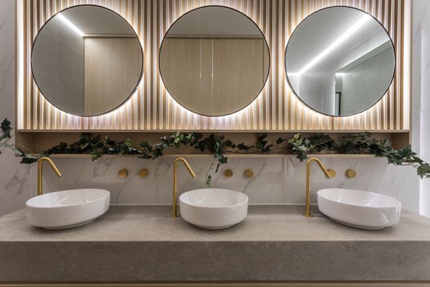 A refined contemporary bathroom vanity with three white vessel basins, brushed gold wall-mounted faucets, and round mirrors set into a vertical timber-slat feature wall. A pale marble-look splashback, grey stone benchtop, integrated under-bench cabinetry, and a planted shelf complete the warm, softly lit fit-out.