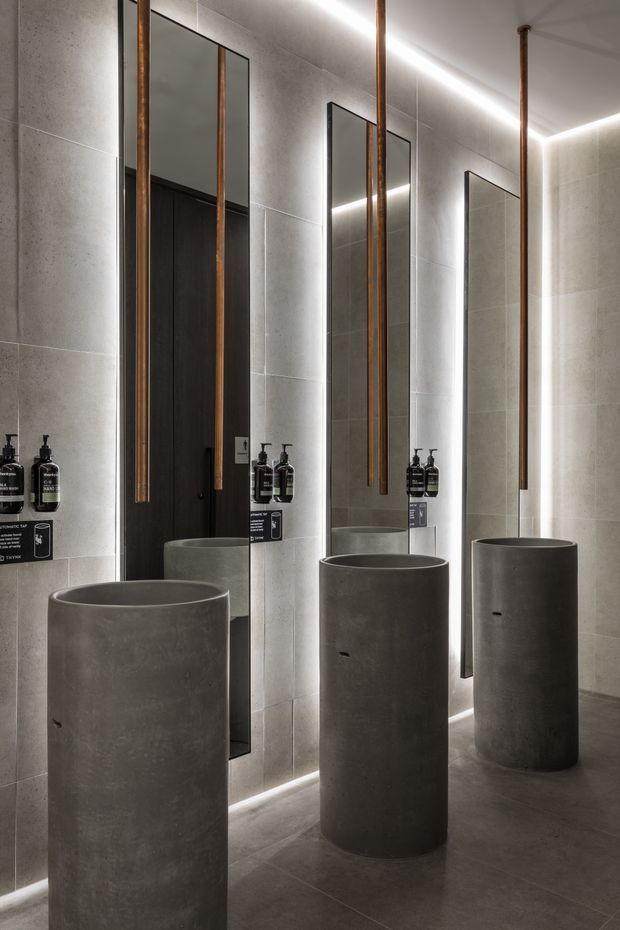 A dark, sophisticated bathroom featuring three cylindrical concrete pedestal basins, large frameless wall mirrors with integrated linear LED lighting, grey stone-look wall and floor tiles, and slender aged copper or bronze wall-mounted tapware. Black doors and dark soap dispensers add contrast.