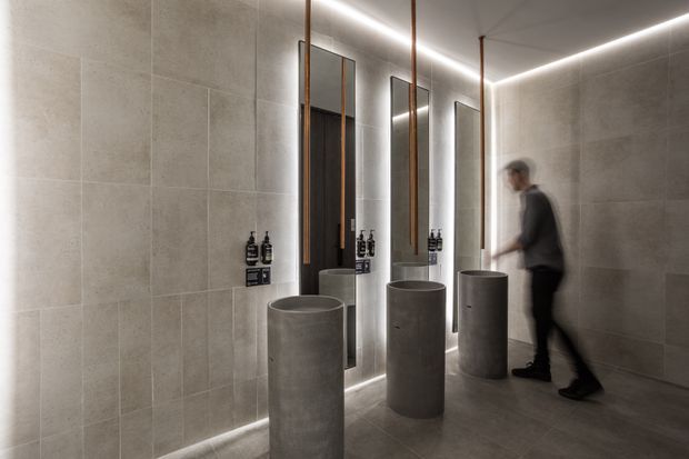 A contemporary bathroom with three cylindrical concrete pedestal basins arranged along a tiled wall. Tall, narrow backlit mirrors with slim bronze-toned frames are mounted above the basins, while integrated linear lighting runs along the ceiling and floor edges. The space has large-format grey stone-look tiles, dark timber door panels, and minimalist black tapware and accessories.