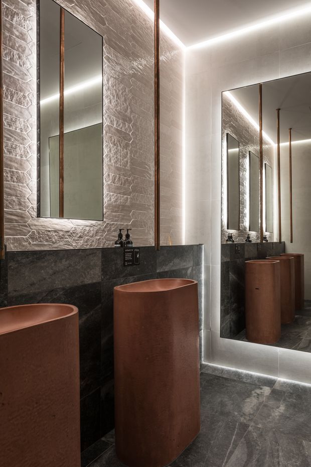 A dramatic contemporary bathroom featuring two tall cylindrical copper-toned vessel basins, dark stone wall cladding and flooring, textured light stone feature walls, slim vertical copper accents, and large illuminated mirrors creating repeated reflections. Integrated linear lighting gives the space a refined, moody atmosphere.
