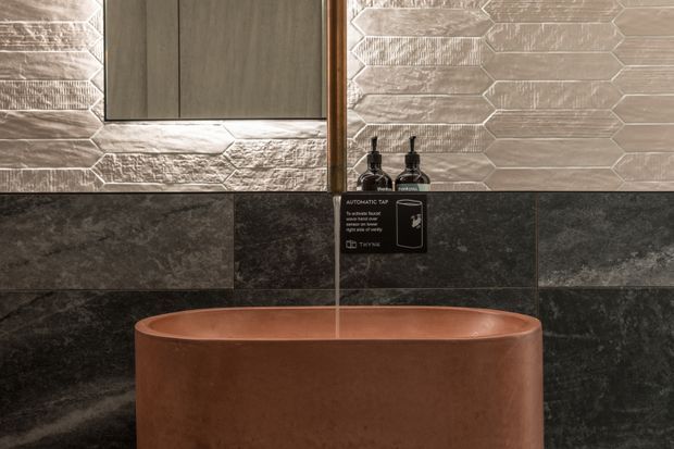 Close-up of a contemporary bathroom vanity area featuring a freestanding copper-toned basin with an integrated automatic tap, dark stone-look wall tiling, textured light stone-look feature tiles, and a mirrored cabinet above.