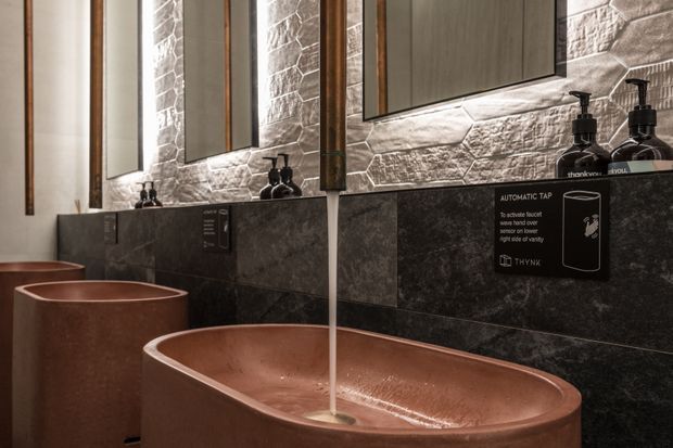 Close-up of a contemporary bathroom vanity featuring sculptural terracotta or copper-toned vessel basins, wall-mounted black tapware, dark stone-look wall tiling, and a textured light stone splashback. Vertical timber-framed mirrors and black soap dispensers complete the warm, refined fit-out.
