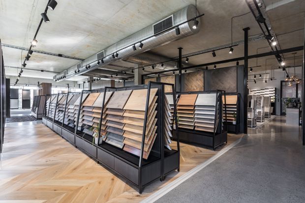 A large, contemporary flooring and materials showroom with rows of black metal display racks presenting timber and tile samples. The space features a herringbone timber floor, polished concrete flooring, exposed concrete ceilings, extensive track lighting, and visible mechanical ductwork.