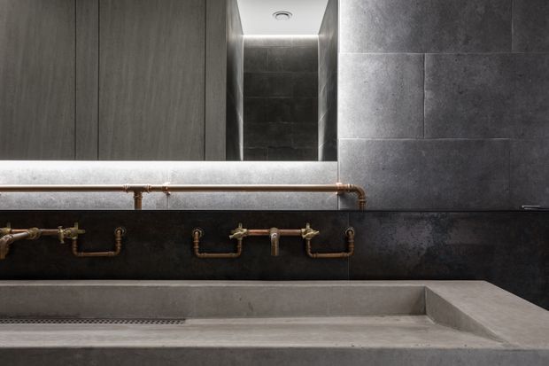 A dark, industrial-style bathroom vanity with a long concrete trough basin, exposed copper plumbing, charcoal wall tiles, dark cabinetry, and a recessed mirrored niche with concealed lighting.