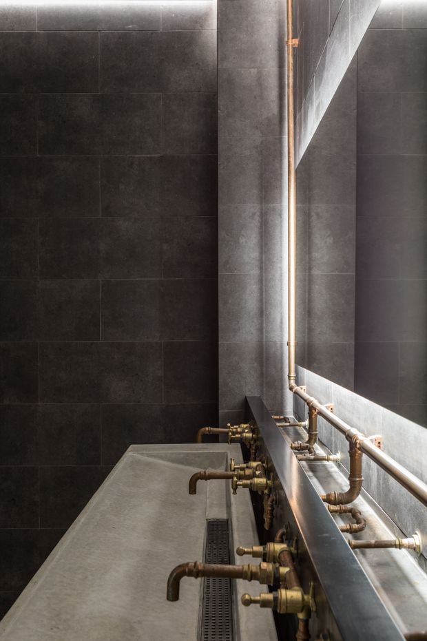 A narrow industrial-style bathroom featuring dark large-format tiled walls, a long concrete trough basin, multiple exposed brass and copper-toned plumbing fixtures, and a large frameless mirror. The space has a moody, utilitarian character with strong linear detailing and minimal decoration.