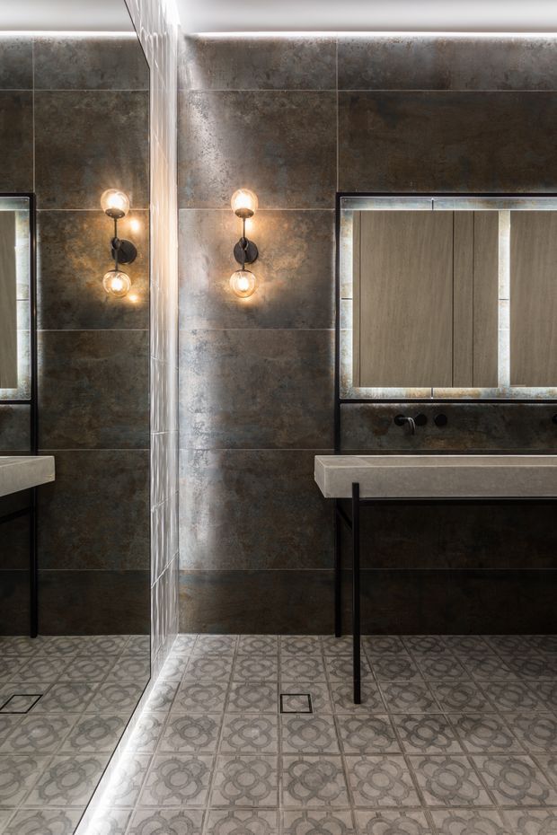 A dark, refined bathroom featuring large mottled charcoal and bronze wall tiles, patterned grey floor tiles, a wall-mounted concrete-look vanity basin with black metal supports, a large framed mirror, and twin globe wall sconces. A frameless glass shower partition is visible at the left.