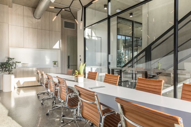 A bright contemporary conference room with a long white meeting table surrounded by tan leather office chairs. The space features full-height black-framed glass partitions, light timber wall panelling, polished concrete flooring, exposed ductwork, and distinctive geometric pendant lighting. A glazed stairwell or adjacent circulation space is visible beyond the partition.