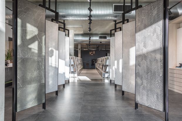 A modern tile and finishes showroom with a central aisle lined by freestanding display panels and material samples. The space features large-format dark grey floor tiles, stone-look and herringbone-patterned wall panels, black metal framing, exposed ductwork, and industrial-style pendant lighting.