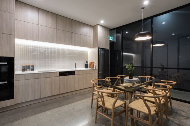 A contemporary open-plan kitchen and dining area with full-height light timber-look cabinetry, a long illuminated tiled splashback, integrated appliances, and a compact island-free layout. A glass dining table with woven timber chairs sits beside black-framed glazed doors or windows, creating a refined evening setting. Recessed ceiling lights and pendant lighting provide warm illumination, while the polished grey floor and dark glazing add contrast.