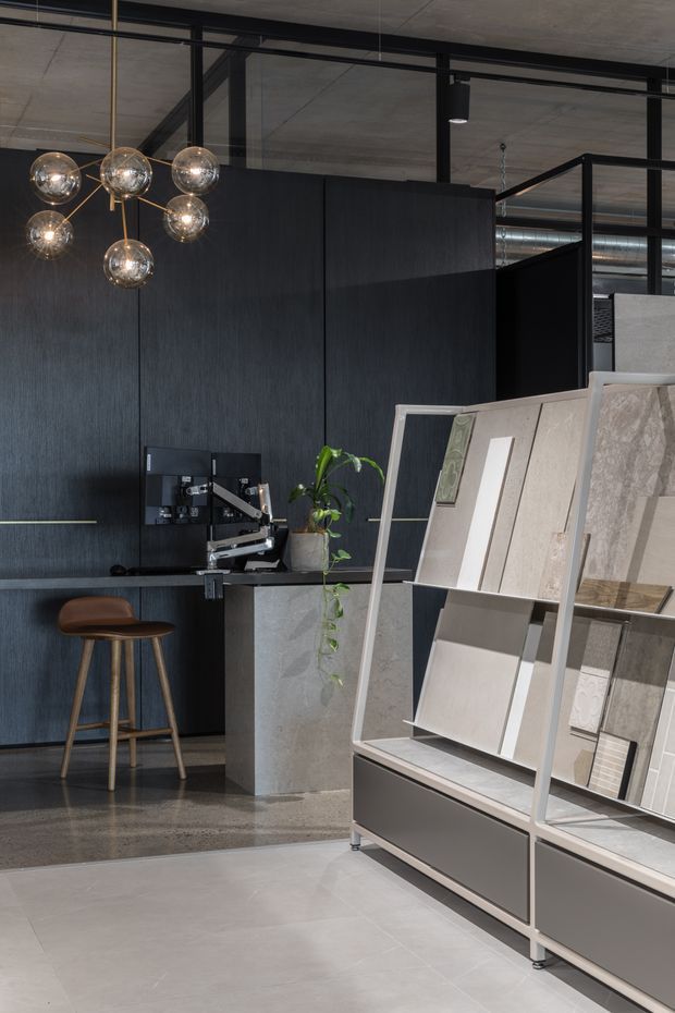 A sophisticated materials showroom or design studio with dark textured wall panelling, exposed concrete ceilings, black-framed glazed partitions, polished concrete flooring, and a display rack presenting large stone or tile samples. A compact workstation with monitor and drafting equipment sits beside a stone-clad counter, accompanied by a timber bar stool, potted plant, and sculptural globe pendant light.