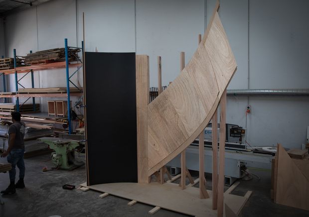 A large-scale timber fabrication workshop showing a curved laminated wood structure mounted on a temporary timber support frame. Shelving with timber stock, woodworking machinery, workbenches, and unfinished panels surround the partially assembled piece in an industrial production environment.