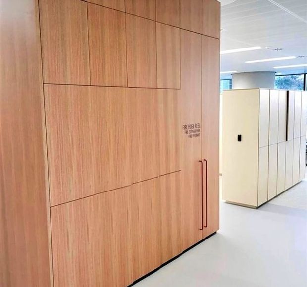 A bright contemporary commercial interior featuring a large full-height timber-faced storage or service wall with flush panel detailing and slim red vertical handles. A small fire-hose-reel notice is printed on the joinery. White built-in cabinetry, pale flooring, a suspended ceiling, and glazed windows are visible in the background.