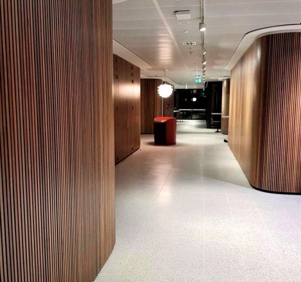 A contemporary commercial interior corridor or lobby with curved walls and extensive vertical timber slat panelling. The space has a pale terrazzo-like floor, a white suspended ceiling with recessed services and track lighting, integrated timber cabinetry, and a small red cylindrical table or reception element. The corridor leads toward a darker rear area and feels refined and softly lit.
